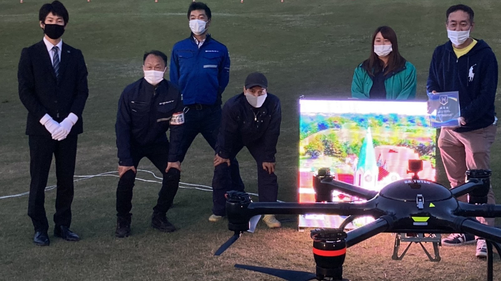 認定楯を持つ人たちと、ドローンで吊り上げた映像モニター(UA日本記録の認定記録ページの写真)